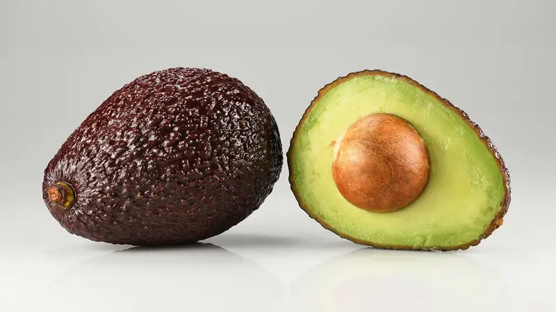 Whole dark purple Hass avocado beside a halved one with pale green flesh and seed