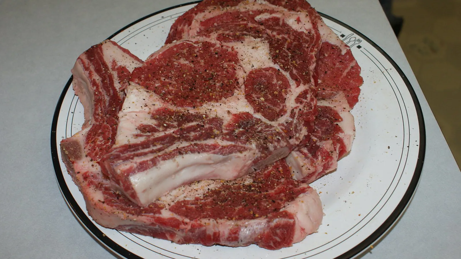 Two seasoned raw bone-in rib steaks on a white plate showing fat cap and marbling