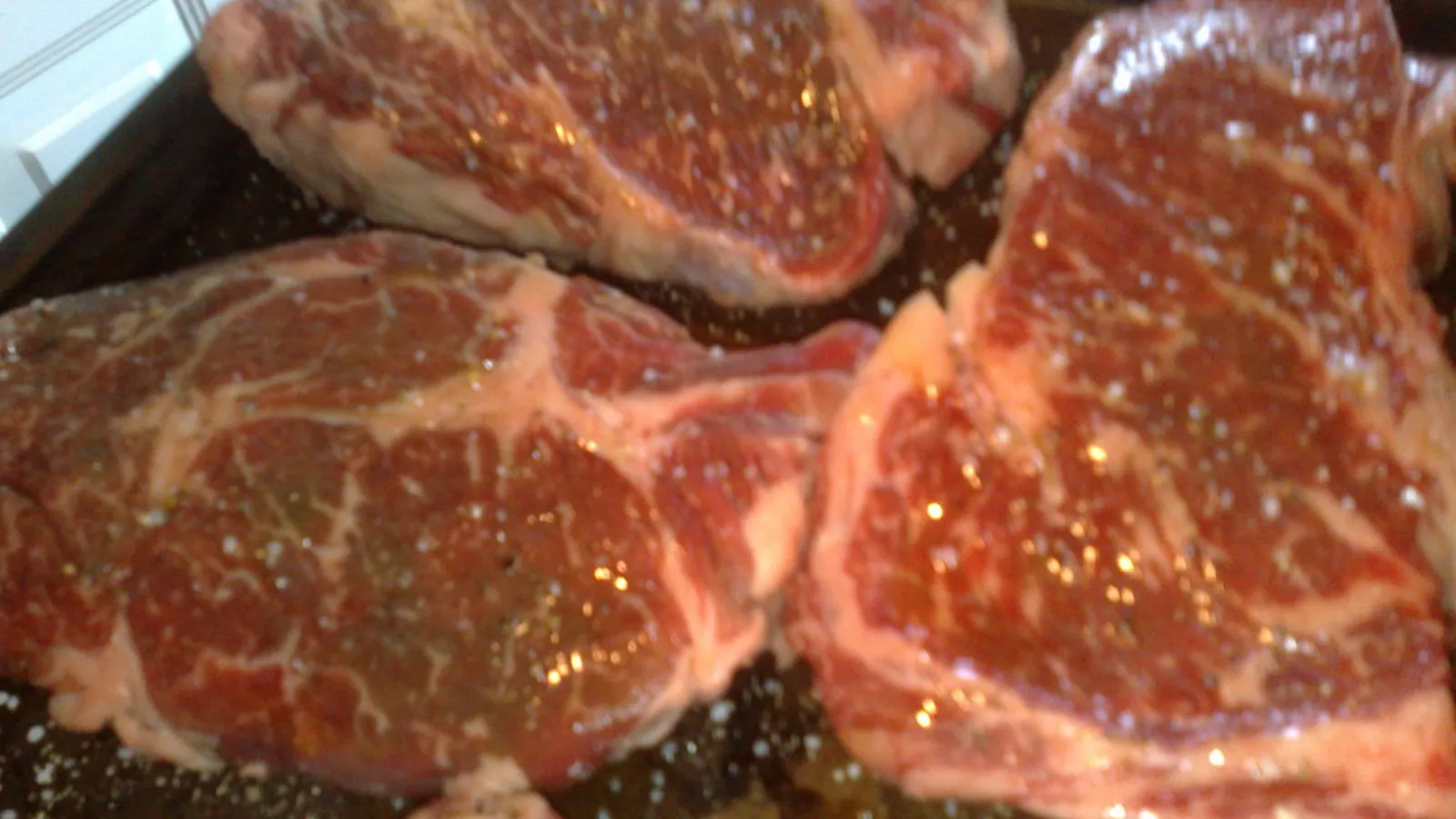 Four raw ribeye steaks with white marbling running through the red meat, sprinkled with salt
