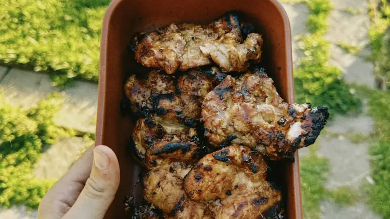 Hand holding a container of grilled bone in chicken thighs with browned, crisp skin