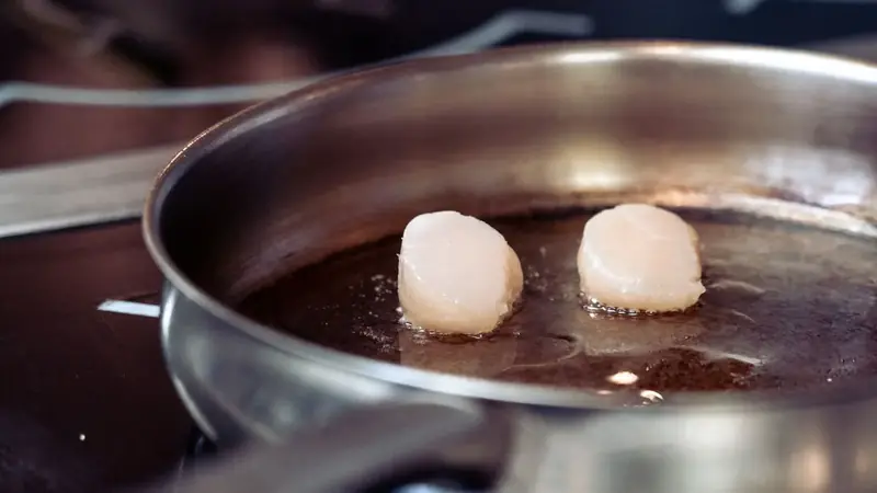 Two scallops searing in a stainless steel pan on a stovetop