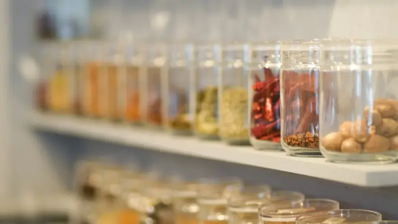 A row of glass jars holding ground spices and whole spices on a kitchen shelf