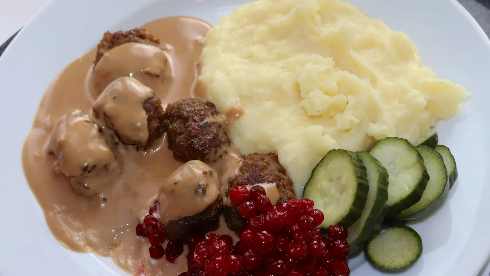 White plate of meatballs with mashed potatoes and sliced cucumbers
