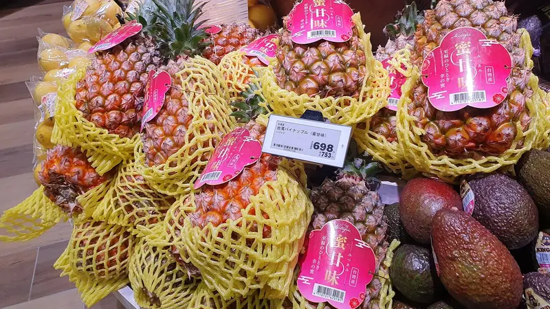 Golden and green pineapples in protective netting on a grocery store display beside avocados