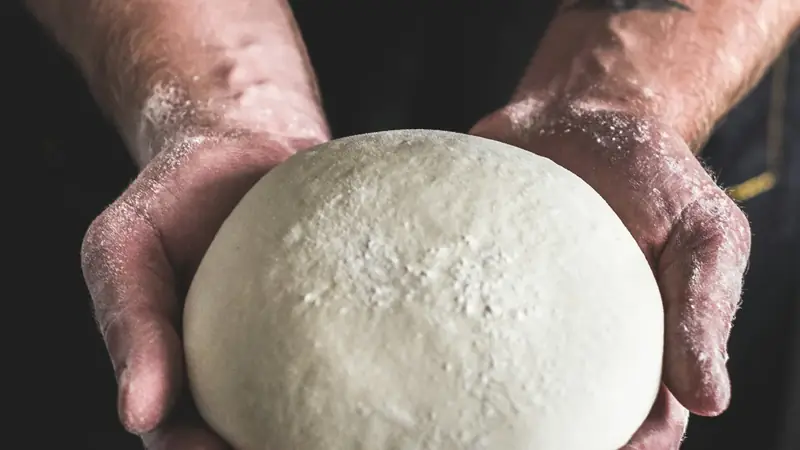 Hands holding a smooth ball of risen pizza dough over a floured surface