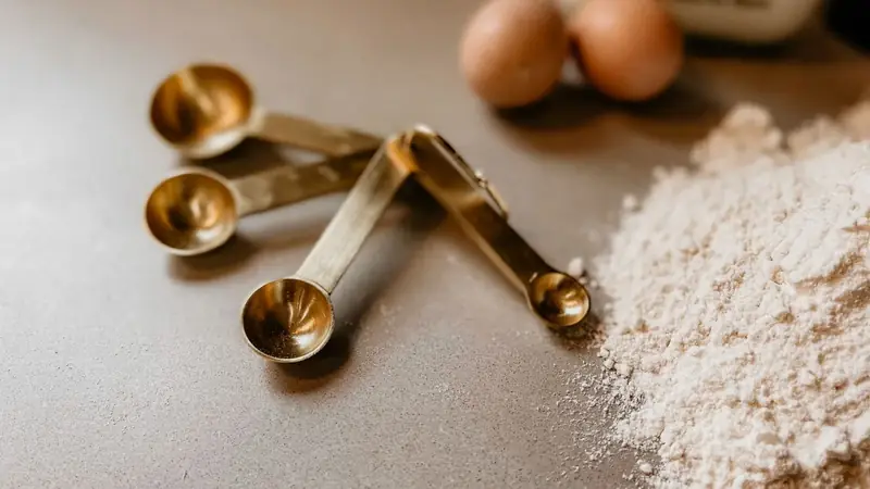 Brass measuring spoons on a kitchen counter beside flour and eggs for baking